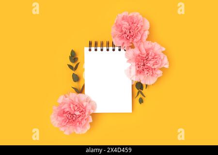 Empty notepad mock up, pink carnation flowers and green eucalyptus branches on a yellow background. Floral composition. Stock Photo