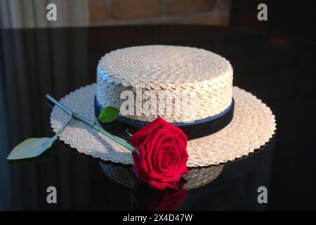 White woven straw hat with a blue ribbon and a red rose on a black background, reflection Stock Photo