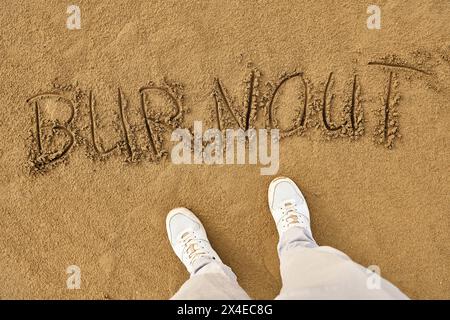 Lido di Jesolo, Italy - 2 May 2024: Burnout, man standing in the sand in front of the lettering, Burnout. Mental health and exhaustion concept *** Burnout, Mann steht im Sand vor dem Schriftzug, Burnout. Mentale Gesundheit und Erschöpfung Konzept Stock Photo