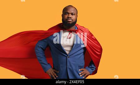 African american superhero flying with red cape, isolated over studio background, flexing muscles. Man wearing cloak posing as hero in costume showing courage and strength, camera B Stock Photo