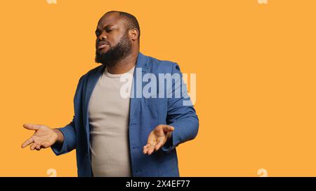 Uncaring man shrugs shoulders, showing indifference and apathy while talking with friend. Careless person doing nonchalant indifferent hand gesturing during conversation, studio background, camera A Stock Photo