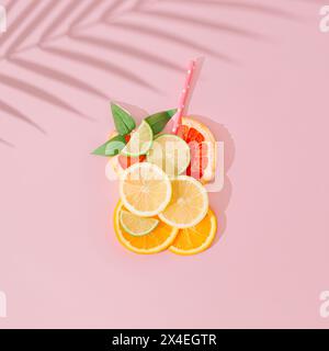 Glass of refreshing drink made of lemon, lime, orange and grapefruit slices, straw and green leaves with palm tree leaf shadow on pink background. Stock Photo