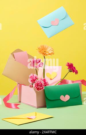 On a yellow background, fresh flowers with gerbera and carnation decorated with cute handmade envelopes on a pink gift box. Creative idea for women’s Stock Photo