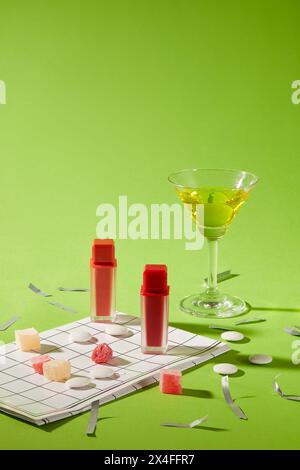 Two lipsticks unbranded displayed with glass cup of wine, jams, go set and silver confetti decorated on a green background. Holiday decorations concep Stock Photo
