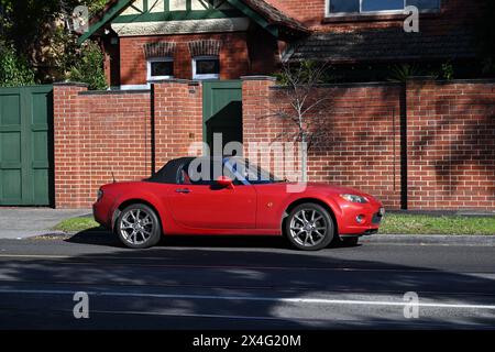 Side view of a bright red Mazda MX-5 convertible, parked in the street outside a suburban home with a red brick wall, during a sunny day Stock Photo