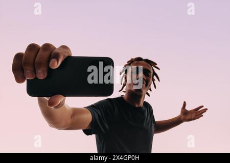 Dreadlocked young man holds a smartphone in a selfie pose against a pink studio backdrop. Gen Z man capturing a self-portrait. Stock Photo