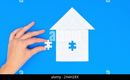 Hand delicately inserting the last piece into a house-shaped puzzle with a roof, set against a serene blue background. Stock Photo