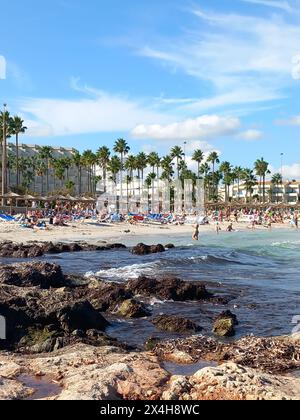 tropical beach oasis: vibrant beachgoers basking in the sun on a lively white sand beach with palm trees, rocky coastline, and modern hotels Stock Photo