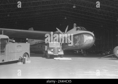 England c.1957 – A photograph of the Dan Air Bristol Type 170 Freighter Mk.31E, registration G-AINL parked within a maintenance hangar. This aircraft, serial number, 012827, was manufactured by The Bristol Aeroplane Company Ltd in 1947. It was obtained by Dan Air in July 1957 from Aviation Traders Ltd, having previously served with the Royal Air Force and Aer Lingus. It was the first of three of its type to go into service with Dan Air. A Bedford O Type flatbed truck is parked under the wing and the airport’s fire service tender is in the foreground. Stock Photo