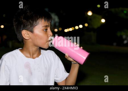 A young Asian boy takes a refreshing drink of water from his bottle while relaxing in a park at nighttime. Stock Photo