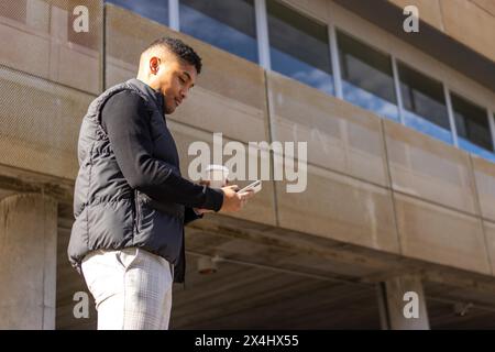 Young businessman walking down the street holding cell phone and glass of coffee, A latin man Walking Down The Street holding glass Coffee And Using Stock Photo