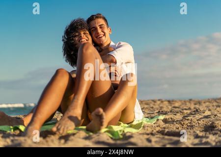 Young interracial couple - laughing and embracing on a sandy beach, a display of affection and joy during a beautiful beach day - lively, romantic. Stock Photo