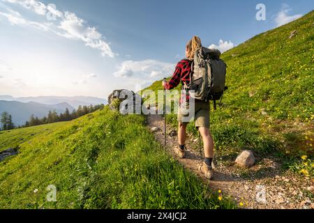 Female hiker with a backpack walking along a gravel mountain trail with lush green slopes and a bright sky at sunset in the german alps. Stock Photo