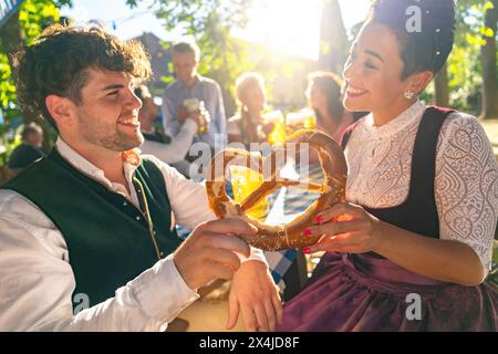 Couple in traditional German tracht holding a pretzel and smiling to each other at oktoberfest or beer garden in bavaria Stock Photo