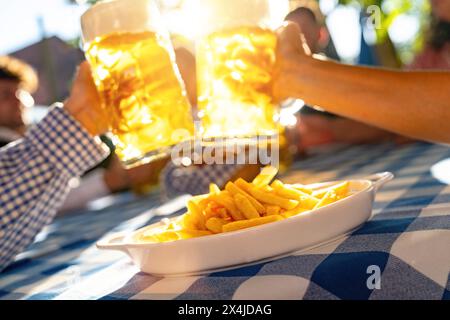 Sunlit beer mugs raised in a toast over a table with fries at Oktoberfest or beer garden in germany Stock Photo