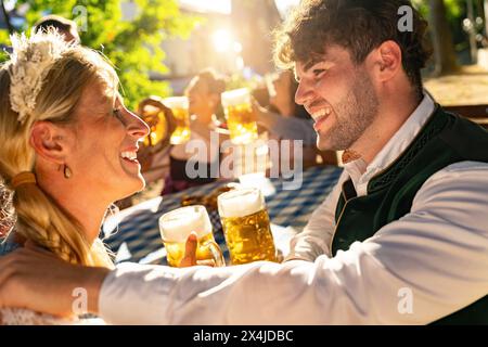 Laughing couple holding beer mugs looking at each other at Oktoberfest or beer garden, with warm sunlight and festive background in germany Stock Photo