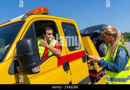 Tow truck operator giving thumbs up to woman with smart phone by damaged car. Roadside assistance concept image. Stock Photo