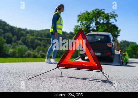 Red warning triangle on road with woman and tow truck in background. Roadside assistance concept image. Stock Photo