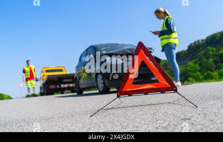 Red warning triangle on road with woman using smartphone and Roadside assistance worker on a tow truck in background. Roadside assistance concept imag Stock Photo
