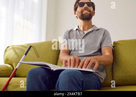 Blind smiling man sitting on sofa at home and reading tactile braille book. Stock Photo