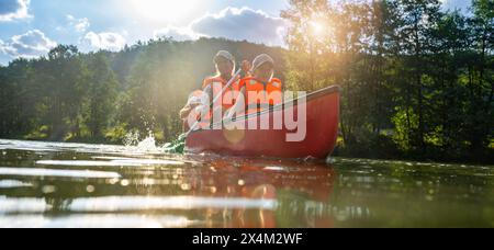 family paddling a red canoe on a river, bathed in sunlight with water splashing around and forest in the background. Family on kayak ride. Wild nature Stock Photo