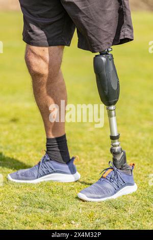 Close up rear view on gray plastic prosthetic leg of single man in red shorts standing in green grass. Close up of a man athlete with a prosthesis on Stock Photo