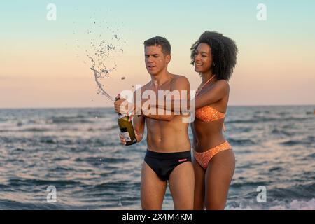 Young interracial couple - celebrating together on the beach at sunset while popping a bottle of sparkling wine, creating joyful and festive moments - Stock Photo