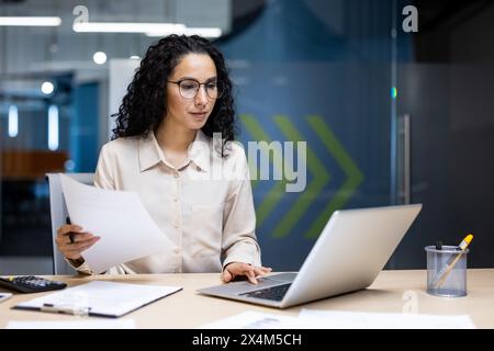 Serious and pensive business woman behind paper work inside office, female financier worker thinks about contracts and reports with charts and graphs, hispanic successful woman uses laptop at work Stock Photo