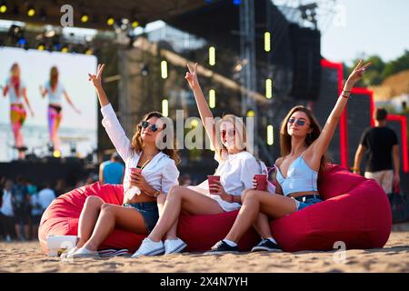 Women celebrate with drinks on beach at sunset. Cheerful ladies sit on bean bags, enjoy cold beverages. Music event, casual outdoor party. Happy group Stock Photo