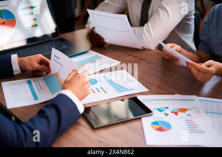 Multiethnic team analyzes business metrics at workplace, discussing colorful graphs on reports, planning strategy. Corporate group collaborates Stock Photo