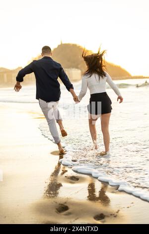 Vertical rear view photo of a caucasian elegant young free young couple running barefoot on the edge of sea water Stock Photo