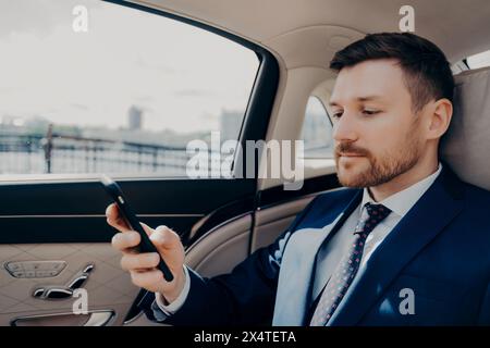 Serene businessman in a luxury car, reviewing information on his smartphone during transit. Stock Photo