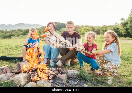 Five kids group Boys and girls cheerfully laughed and roasted sausages on sticks over a campfire flame near the green tent. Outdoor active time spendi Stock Photo