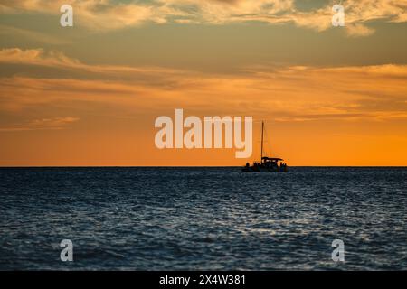 A boat is sailing in the ocean at sunset. The sky is orange and the water is calm Stock Photo