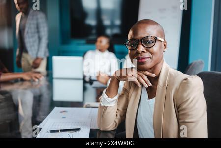 African businesswoman wearing glasses confidently leads a meeting in a boardroom with her colleagues while they discuss documents and reports. Stock Photo