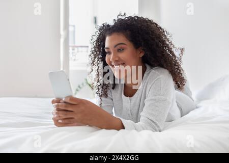 Young Woman Smiling While Using Smartphone in Bright Bedroom Setting Stock Photo