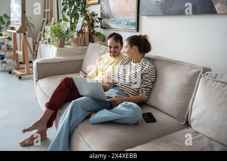 Happy relaxed creative couple of artist smiling sitting with laptop in art studio talking, planning. Stock Photo