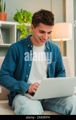 Vertical. Happy freelancer man working at home with a laptop on a sofa. Young guy smiling and browsing on internet using a computer at living room. Entrepreneur looking bank finances and invests. High quality photo Stock Photo