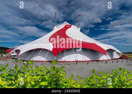 White circus marquee tent with red band on blue sky with white clouds with green hedge in foreground Stock Photo