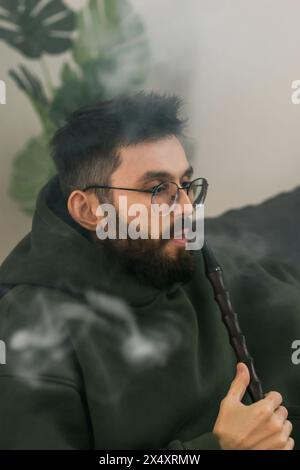 Bearded man is smoking hookah at home and blowing cloud of smoke - chill time and resting Stock Photo