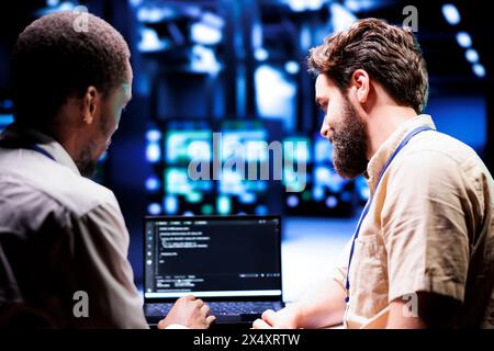 IT developers collaborating in server farm writing script code on laptop terminals. Programmers team conducting maintenance tasks in highly advanced data center, ensuring racks software is up to date Stock Photo