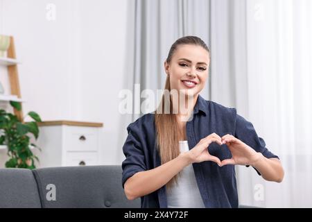 Happy woman showing heart gesture with hands at home, space for text Stock Photo
