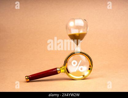 Tax time deadline concept. Percentage icon in magnifying glass lens on hourglass, isolated on brown recycled paper background, minimal style. Interest Stock Photo