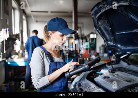 Female auto mechanic using diagnostic tool, scanner, running diagnostic on car. Beautiful woman working in a garage, wearing blue coveralls. Stock Photo