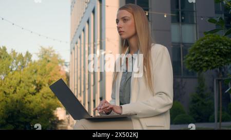Pensive Caucasian woman businesswoman student female business lady girl developer think typing on laptop outdoors street near office work remote Stock Photo
