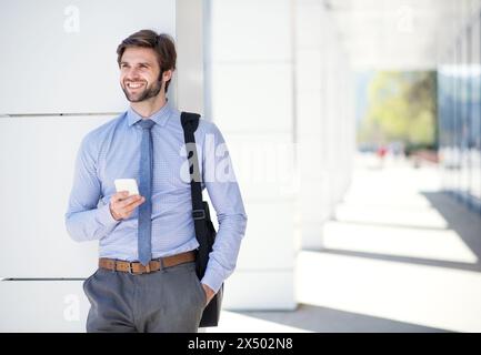 Handsome businessman scrolling on smartphone while walking on city street, going to office. Manager smiling, outdoor in urban setting Stock Photo
