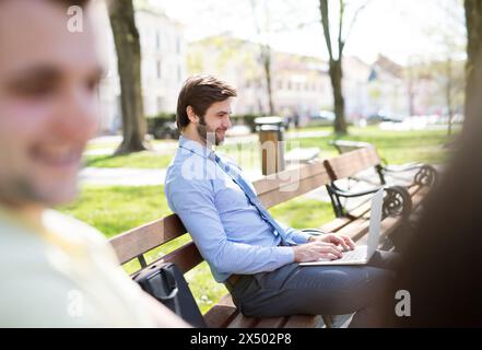 Handsome businessman sitting on city bench, typing on laptop. Working remotely from city park, waiting for business meeting. Manager smiling, outdoor Stock Photo