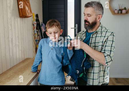 Father helping son get ready for kindergarten, preschool. Putting backpack on his back. Stock Photo