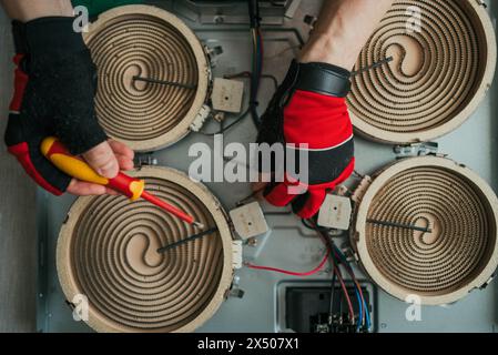 Hands of technician are shown in close-up, repairing and adjusting electrical components inside an electric stove, demonstrating skill and precision. Stock Photo