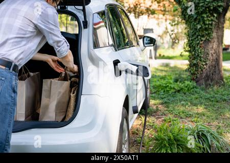 Man with shopping bag next to a charging electric car in the yard of a country house. Stock Photo